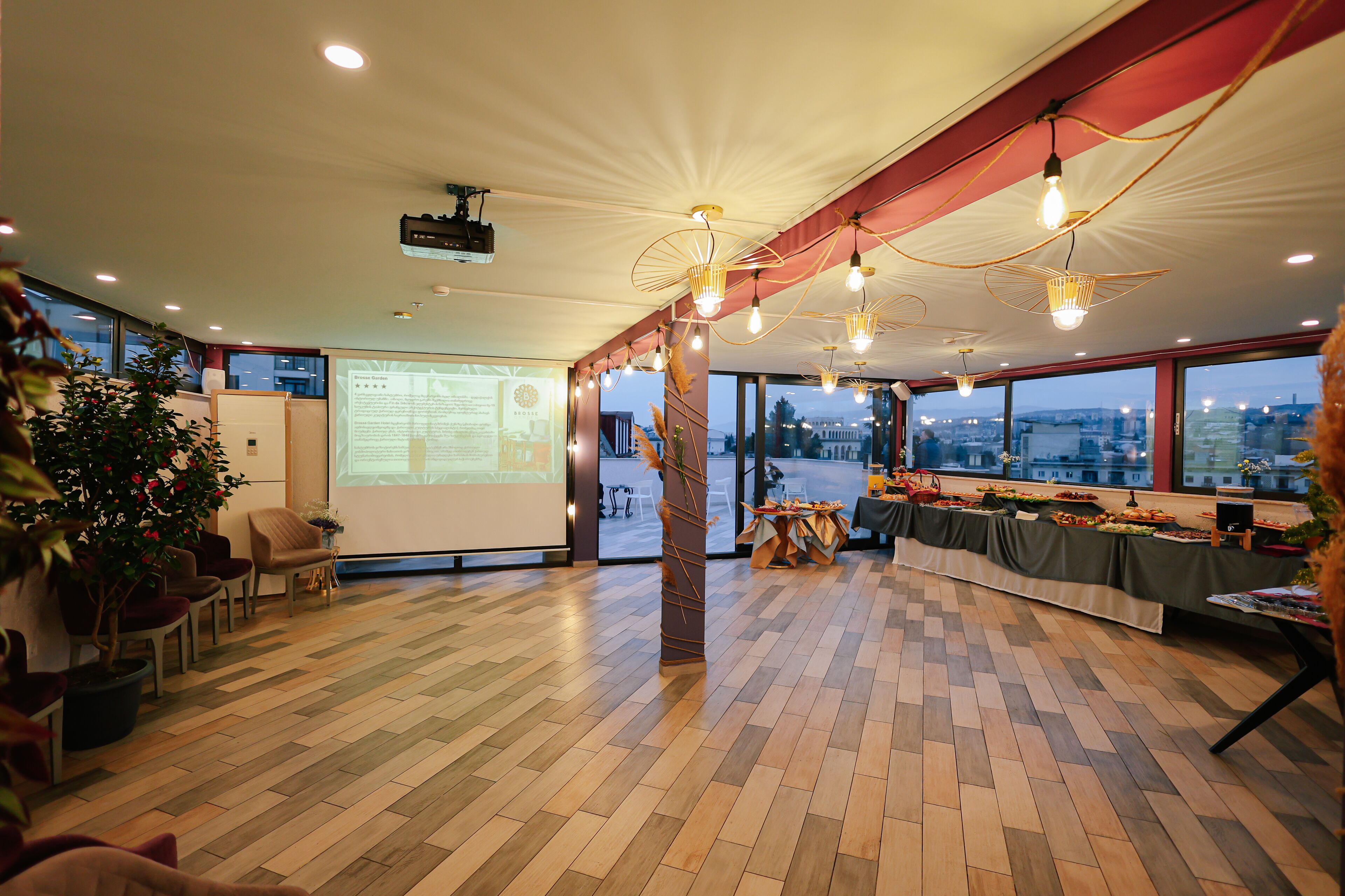 A spacious conference room with a large screen, wooden flooring, and a view of the cityscape through large windows.