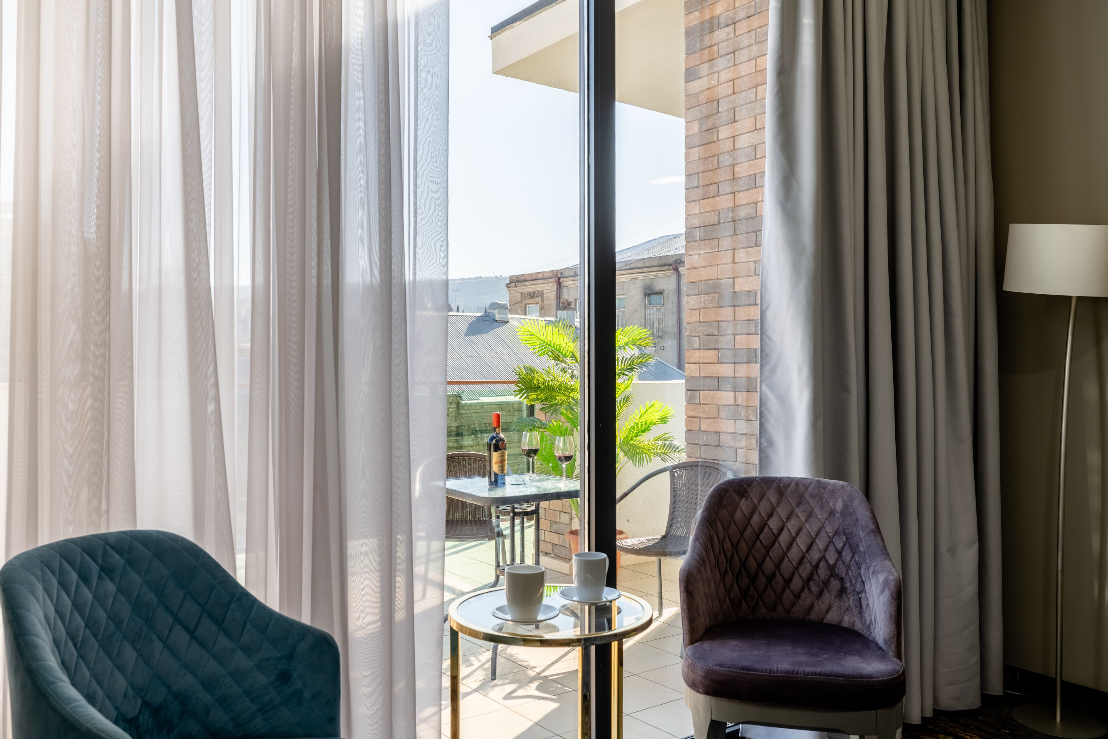 A room with two chairs, a table with a bottle and glasses, and a view of a building through sheer curtains.