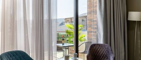 A room with two chairs, a table with a bottle and glasses, and a view of a building through sheer curtains.