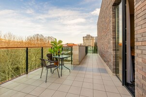 Un balcón con una mesa y sillas, una planta en maceta y vistas a árboles y edificios.