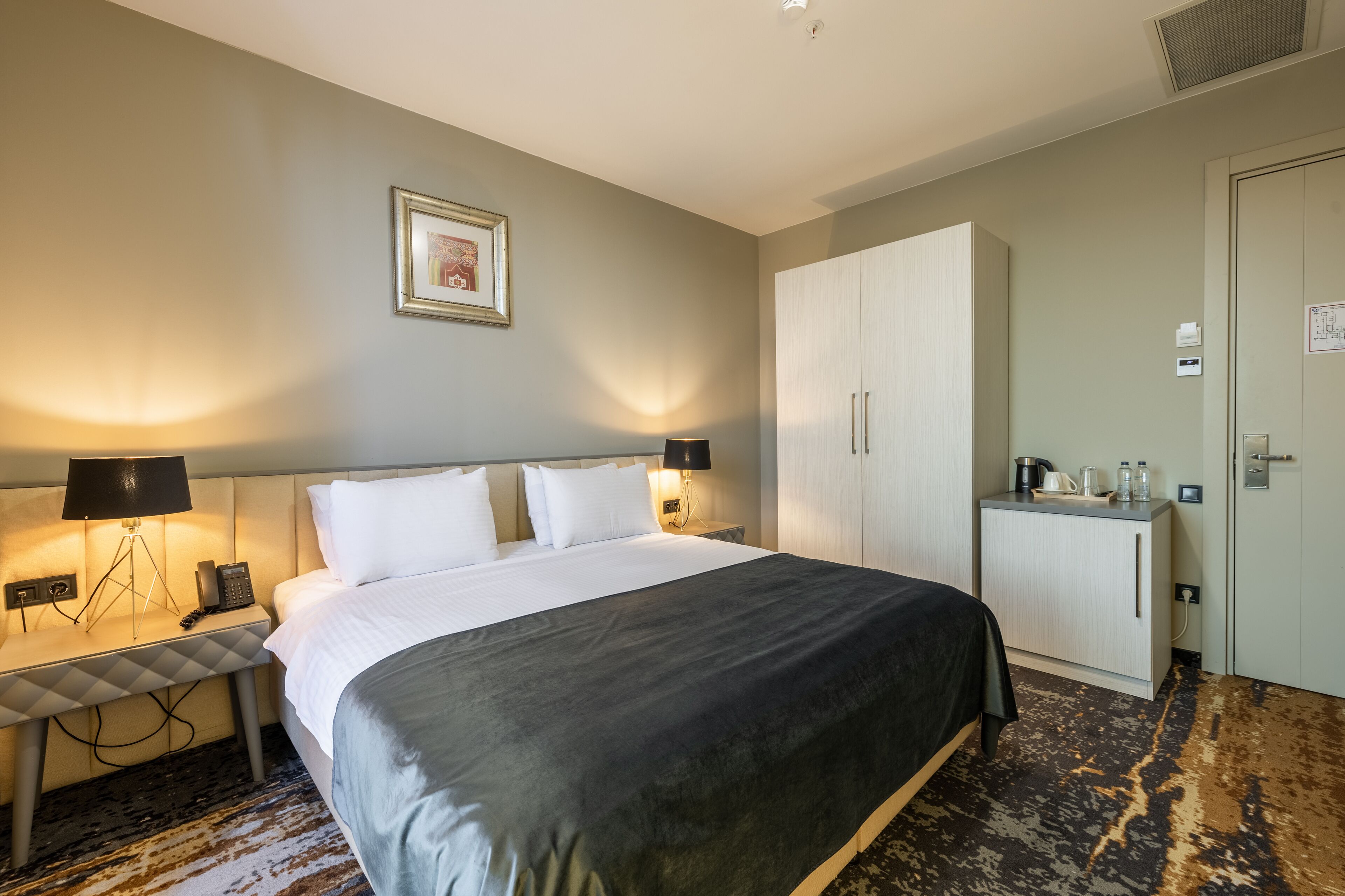 Une chambre d’hôtel avec un grand lit, des lampes de chevet, un bureau avec un téléphone, une armoire et une petite kitchenette.