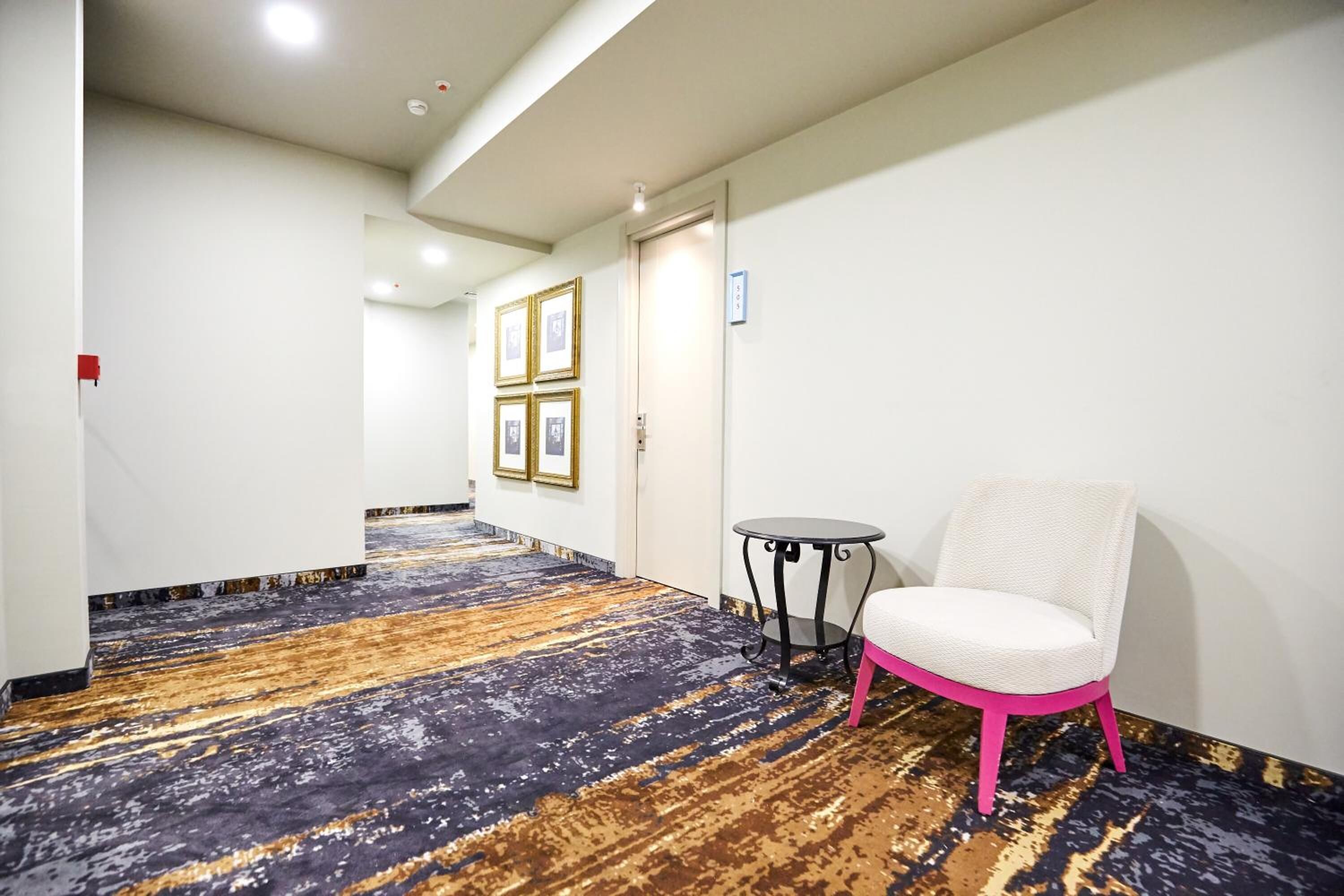 Um corredor de hotel com carpete estampado, uma cadeira branca e uma mesa pequena.