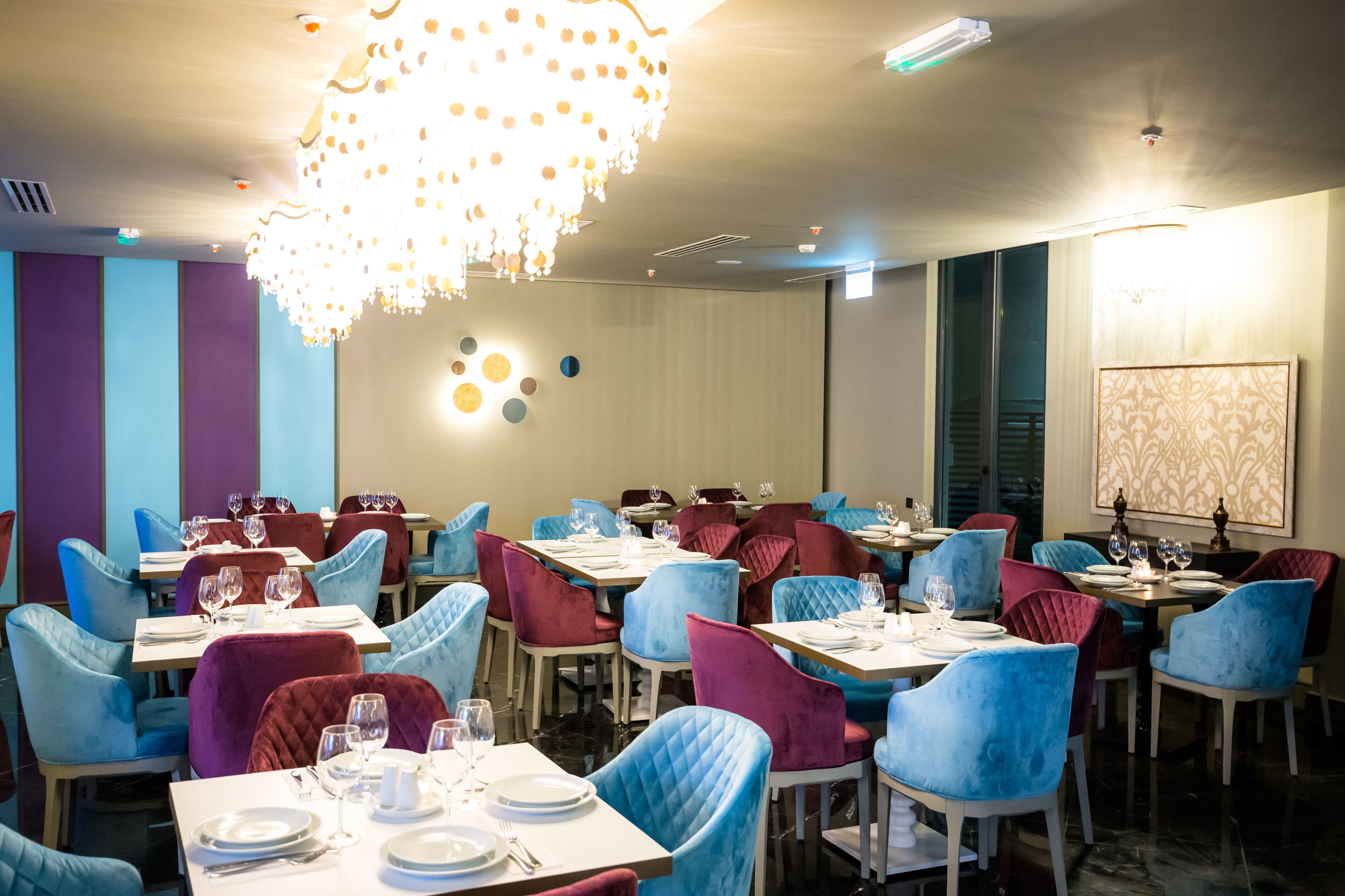 A dining area with tables set for four, blue and red chairs, and a chandelier.