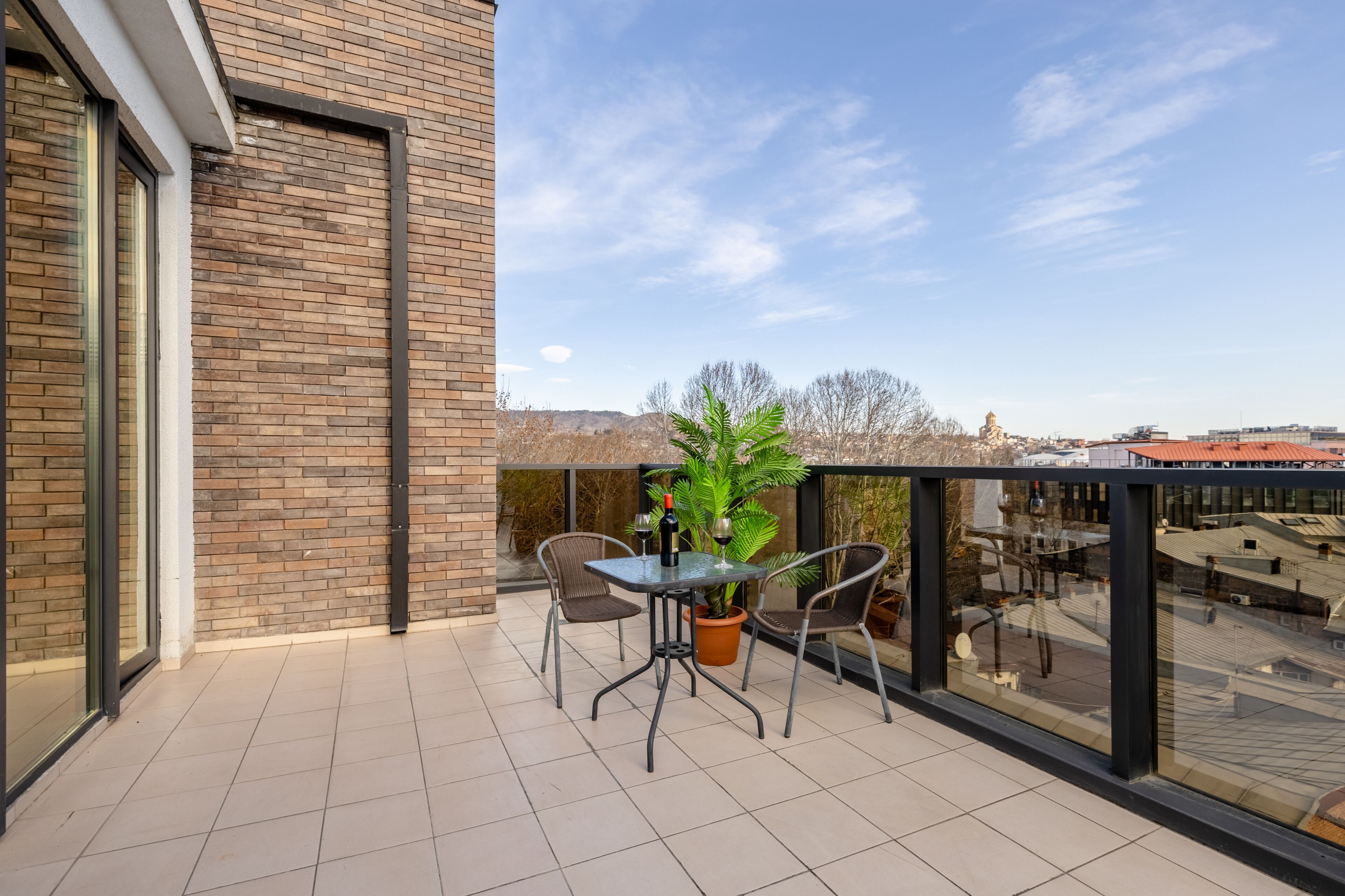 A balcony with a table and chairs, a potted plant, and a bottle of wine.