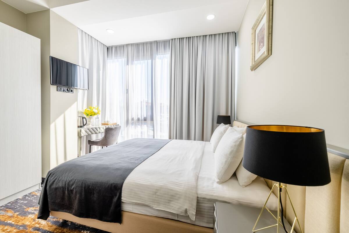 A hotel room with a bed, a desk with a chair, a TV mounted on the wall, and a window with curtains.