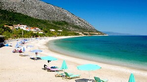 A beach with sun loungers and umbrellas, a clear blue sea, and houses on a hillside. - Faro Del Porto (Kefalonia)