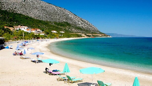 A beach with sun loungers and umbrellas, a clear blue sea, and houses on a hillside. - Faro Del Porto (Kefalonia)
