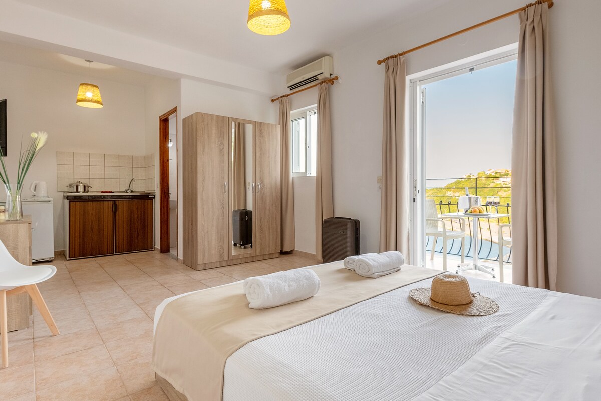 A hotel room with a bed, a desk, a chair, a kitchenette, and a balcony with a view.