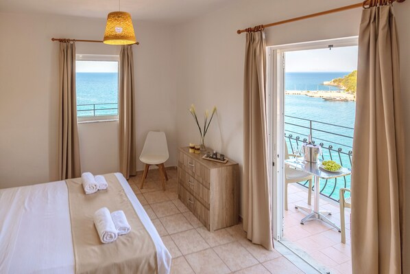 A hotel room with a bed, a desk, a chair, a dresser, and a balcony with a view of the sea. - Faro Del Porto (Kefalonia)