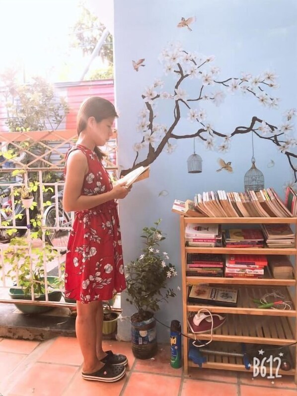 Una persona leyendo un libro en una habitación con una estantería, una planta en maceta y un mural de un árbol con pájaros.