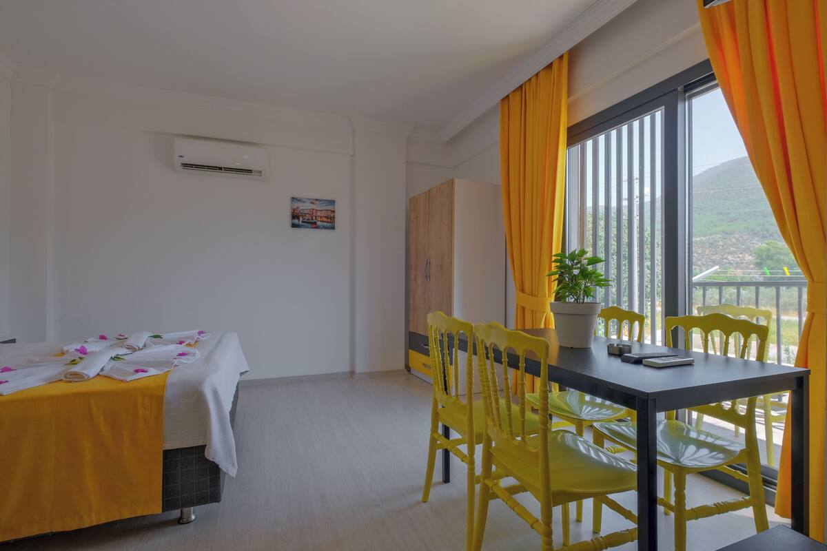 A room with a bed, a table with yellow chairs, a potted plant, and a window with curtains.