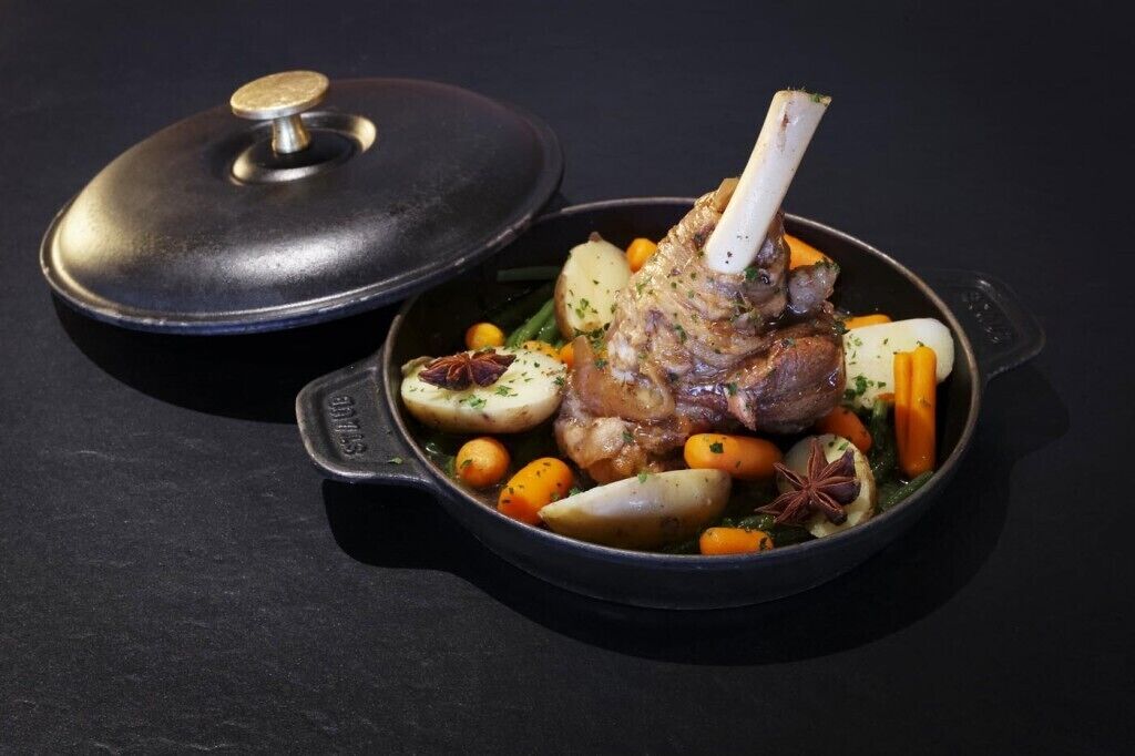 A cast iron skillet with a lamb shank, carrots, potatoes, and herbs.
