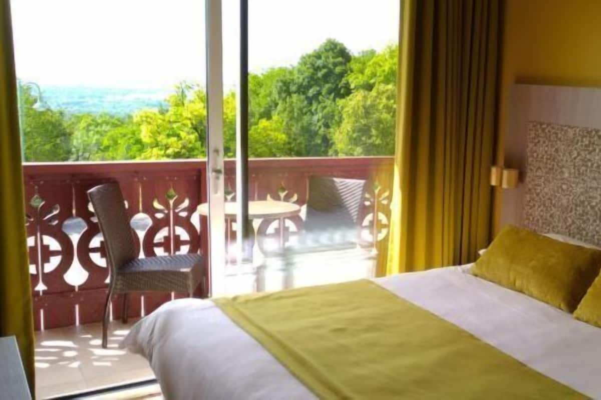 Quarto de hotel com cama, vista para área verde e varanda com cadeiras.