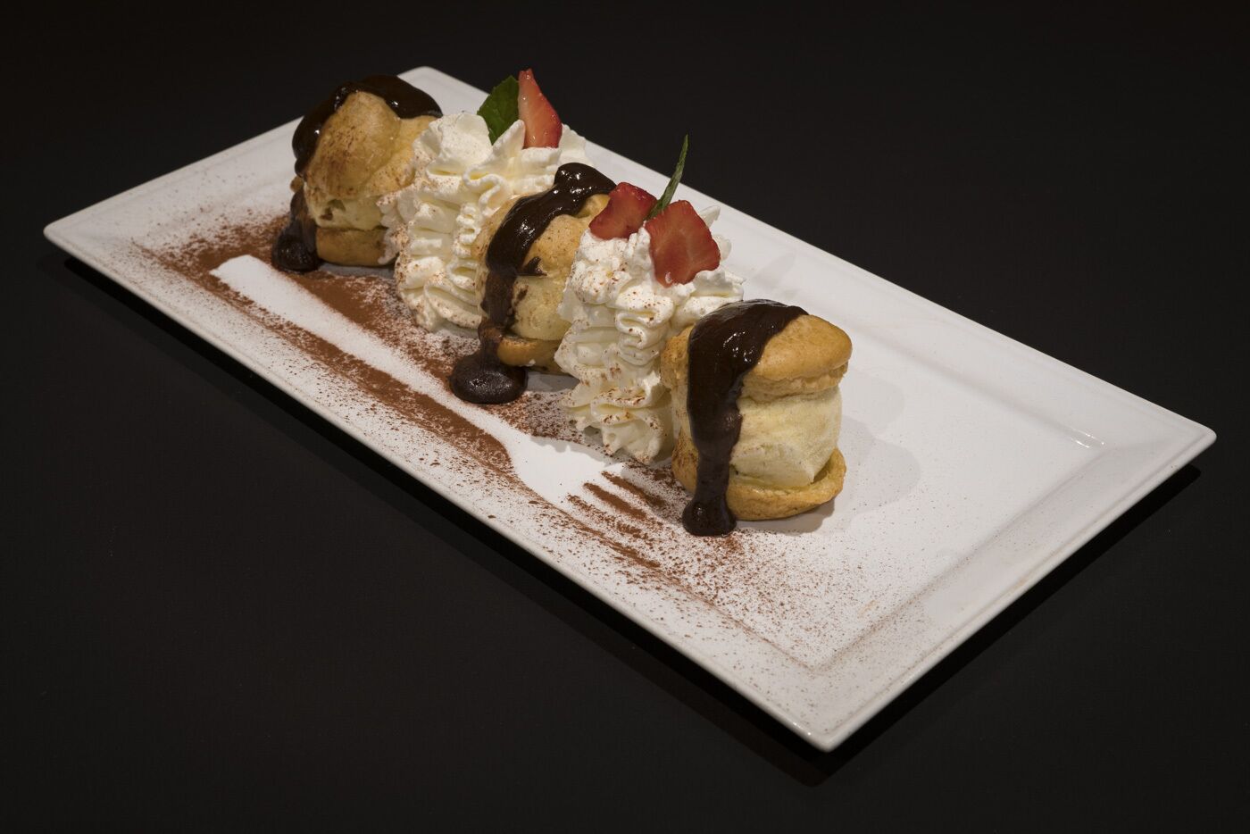 Un postre en un plato rectangular con salsa de chocolate, crema batida y fresas.