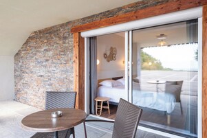 Habitación con cama, una mesita y una silla, con vista a un estacionamiento a través de un ventanal.