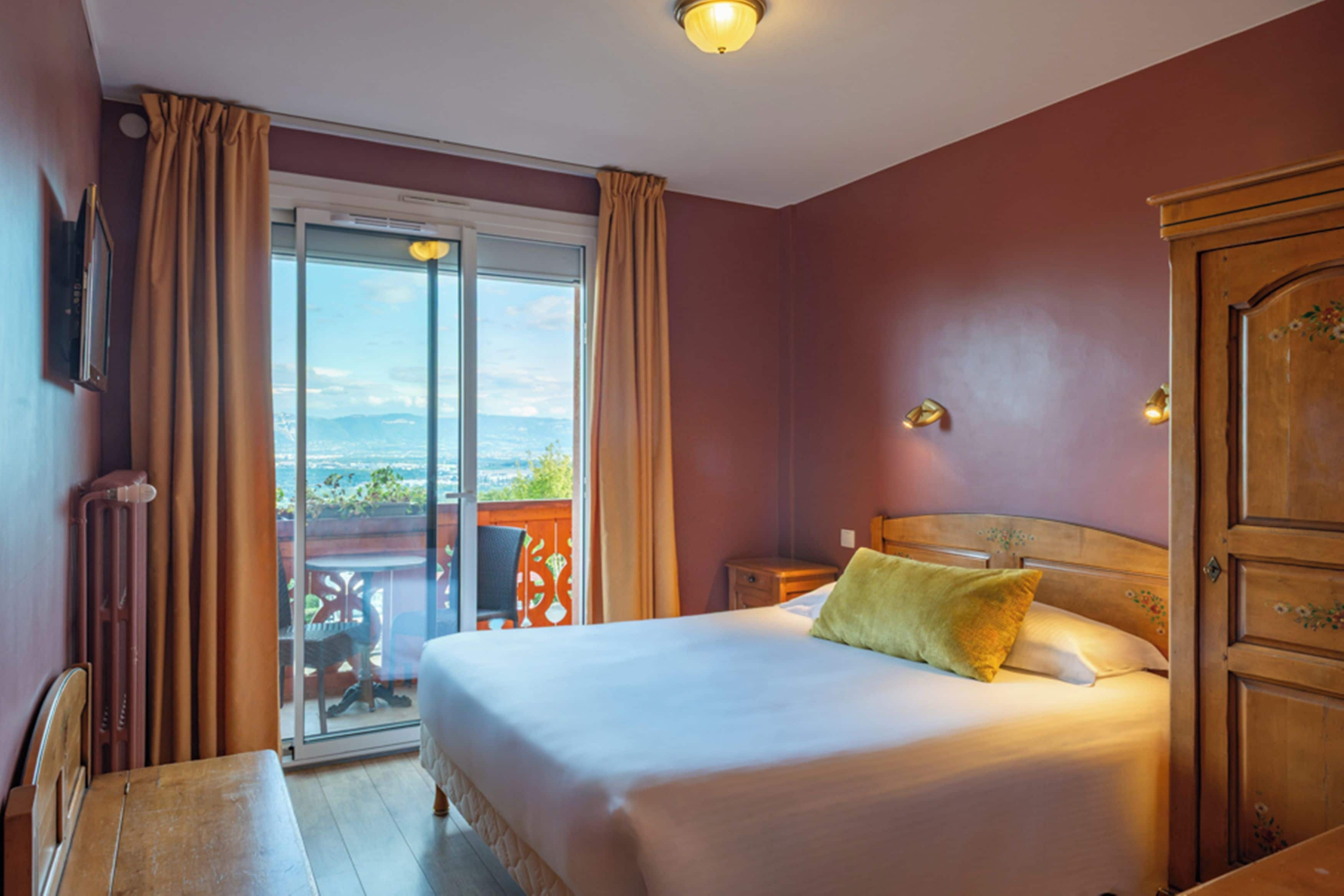 A hotel room with a bed, a nightstand, a wooden wardrobe, a window with curtains, and a view of the outdoors.