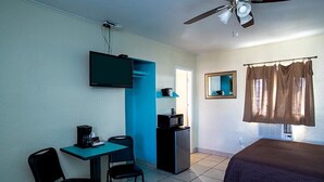 Une petite pièce, de taille modeste, comprenant un lit, un bureau avec une chaise, un téléviseur, un four à micro-ondes et un ventilateur de plafond.