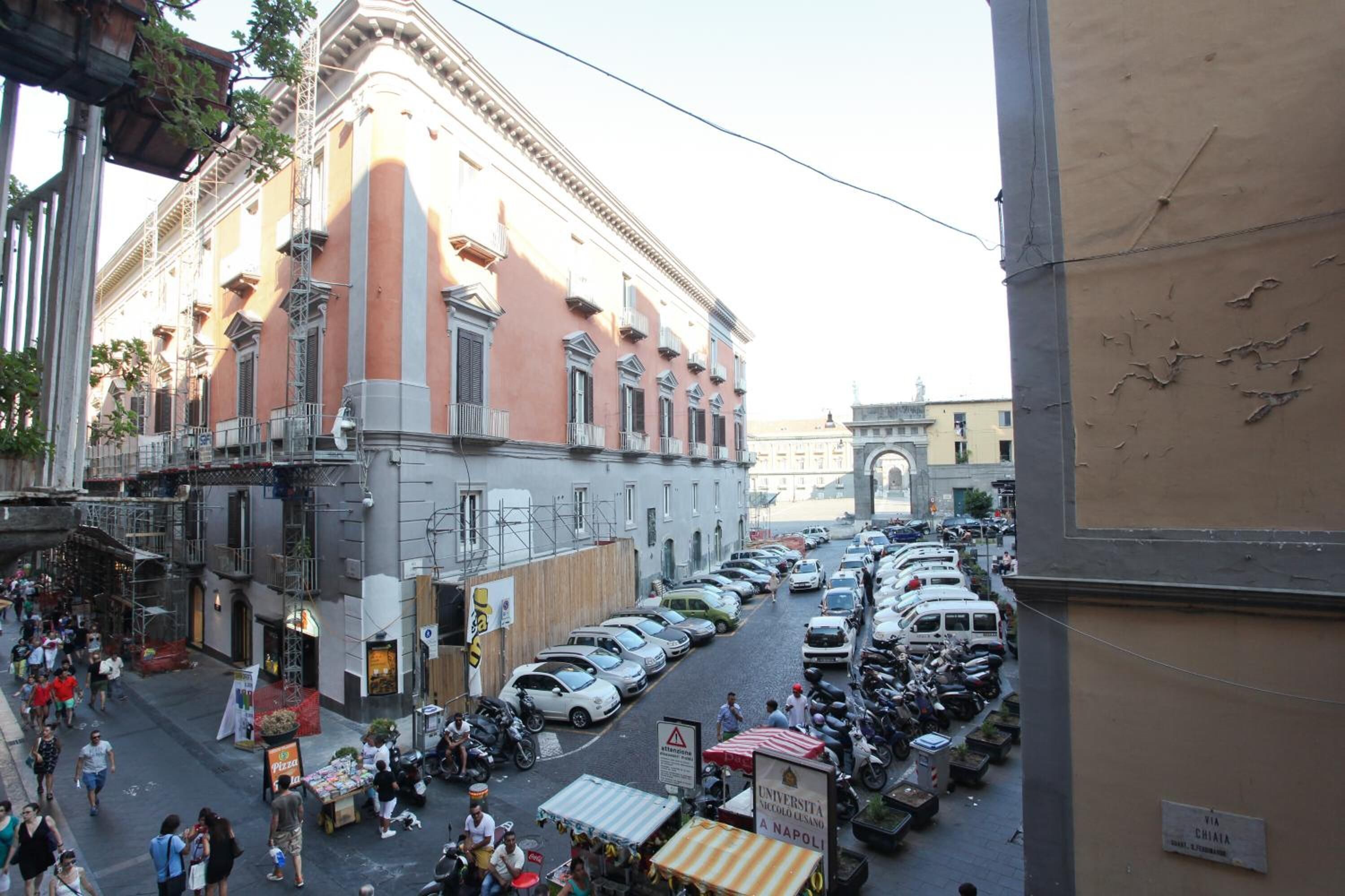 A busy street with parked cars and motorcycles, a building with balconies, and a sign for 'Università Scuola Normale A Napoli'.