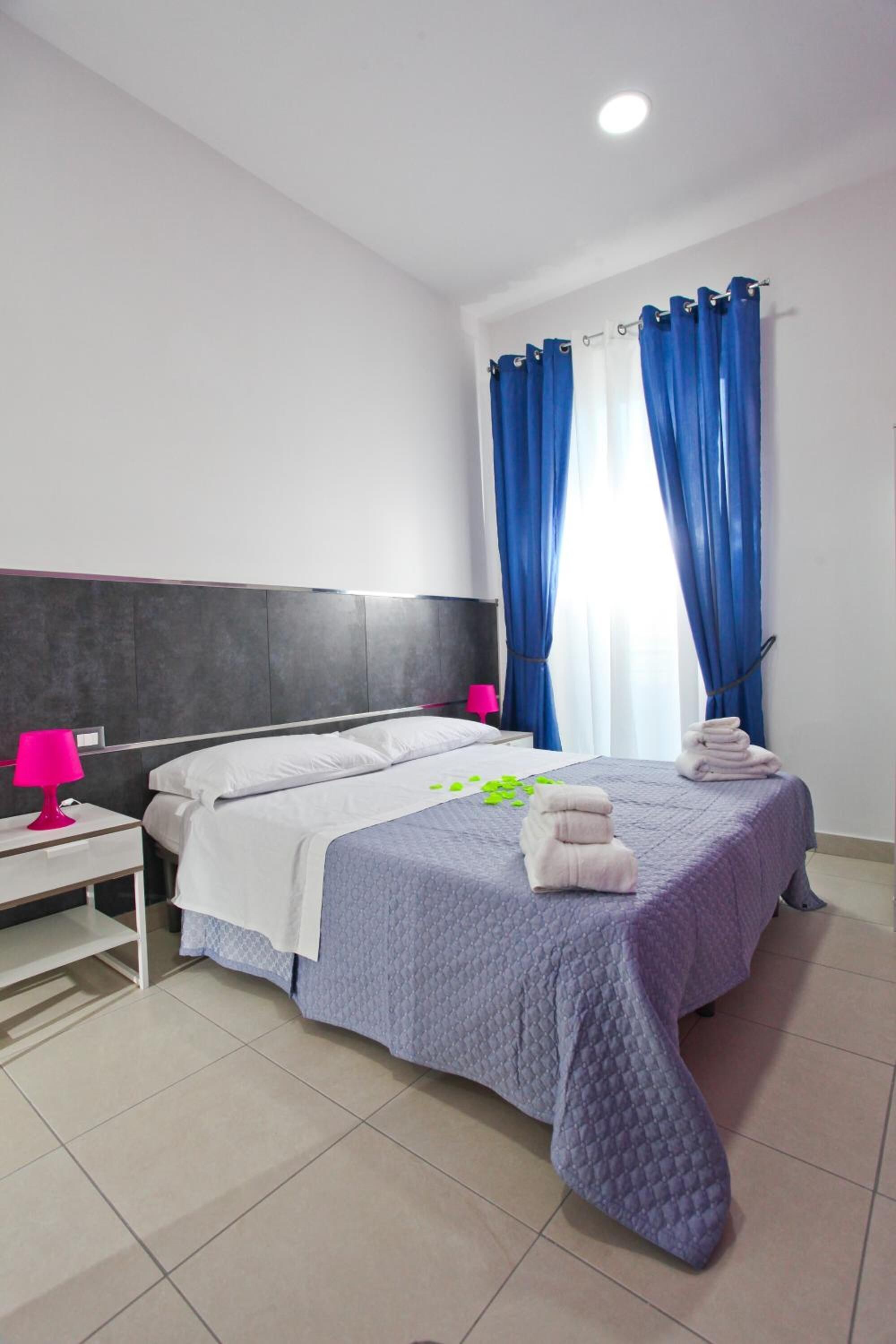 Camera d'albergo con un letto, due comodini, una testiera e una finestra con tende blu.