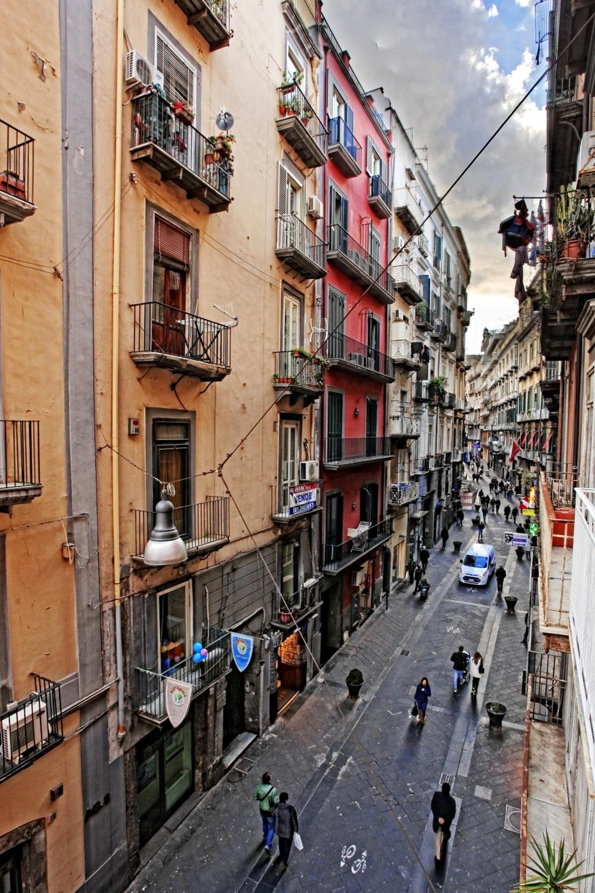 Una calle estrecha flanqueada por edificios de varios pisos, algunos con balcones y plantas en macetas.