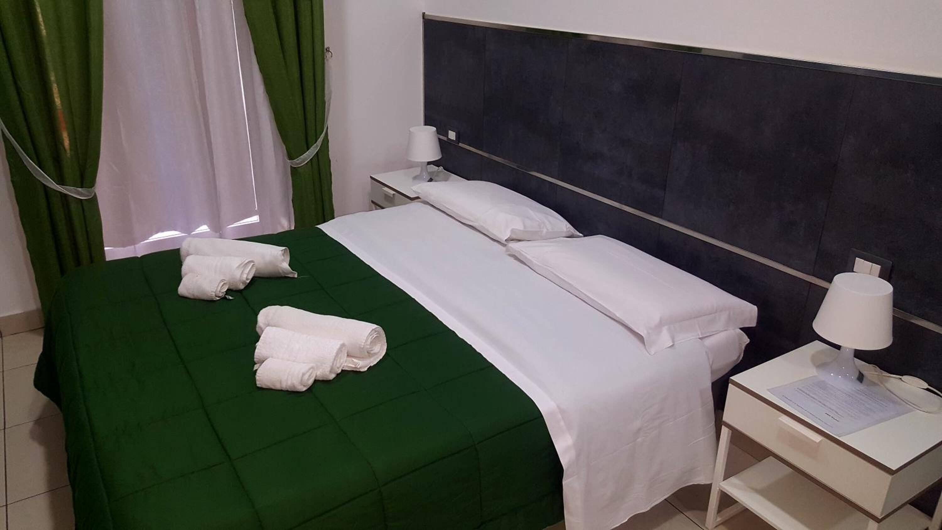 Ein Hotelzimmer mit grüner Bettdecke, weißen Leinentüchern, einem dunklen Kopfteil, einem Nachttisch mit Lampe und einem Vorhang.