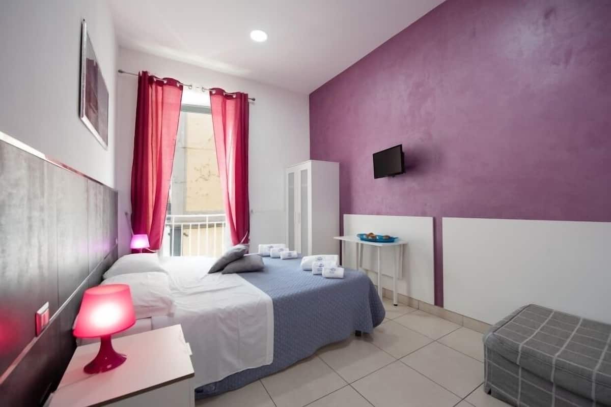 A hotel room with a bed, a nightstand with a lamp, a small table with a tray of items, a TV mounted on the wall, and a window with red curtains.