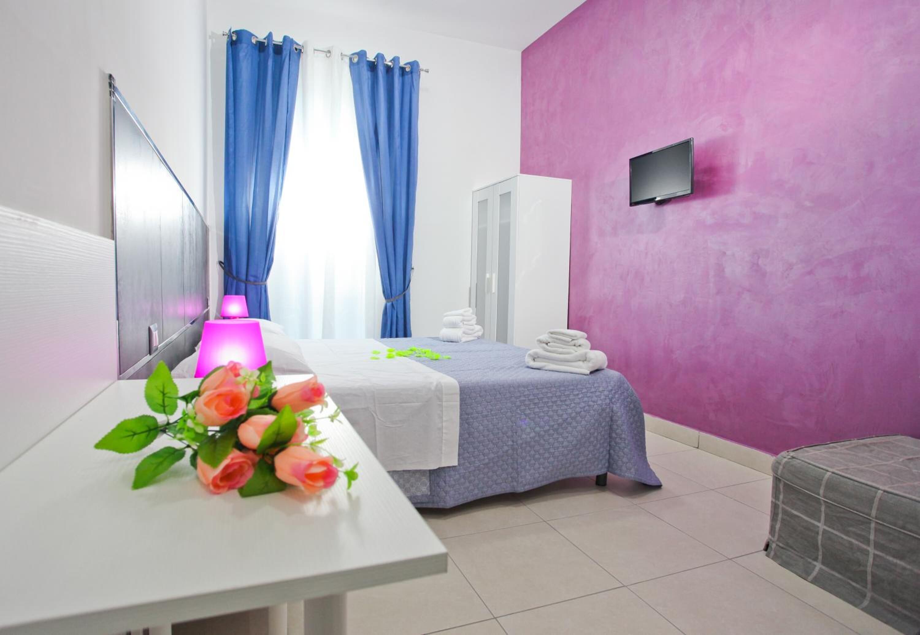 A hotel room with a bed, a desk with a lamp, and a pink wall.