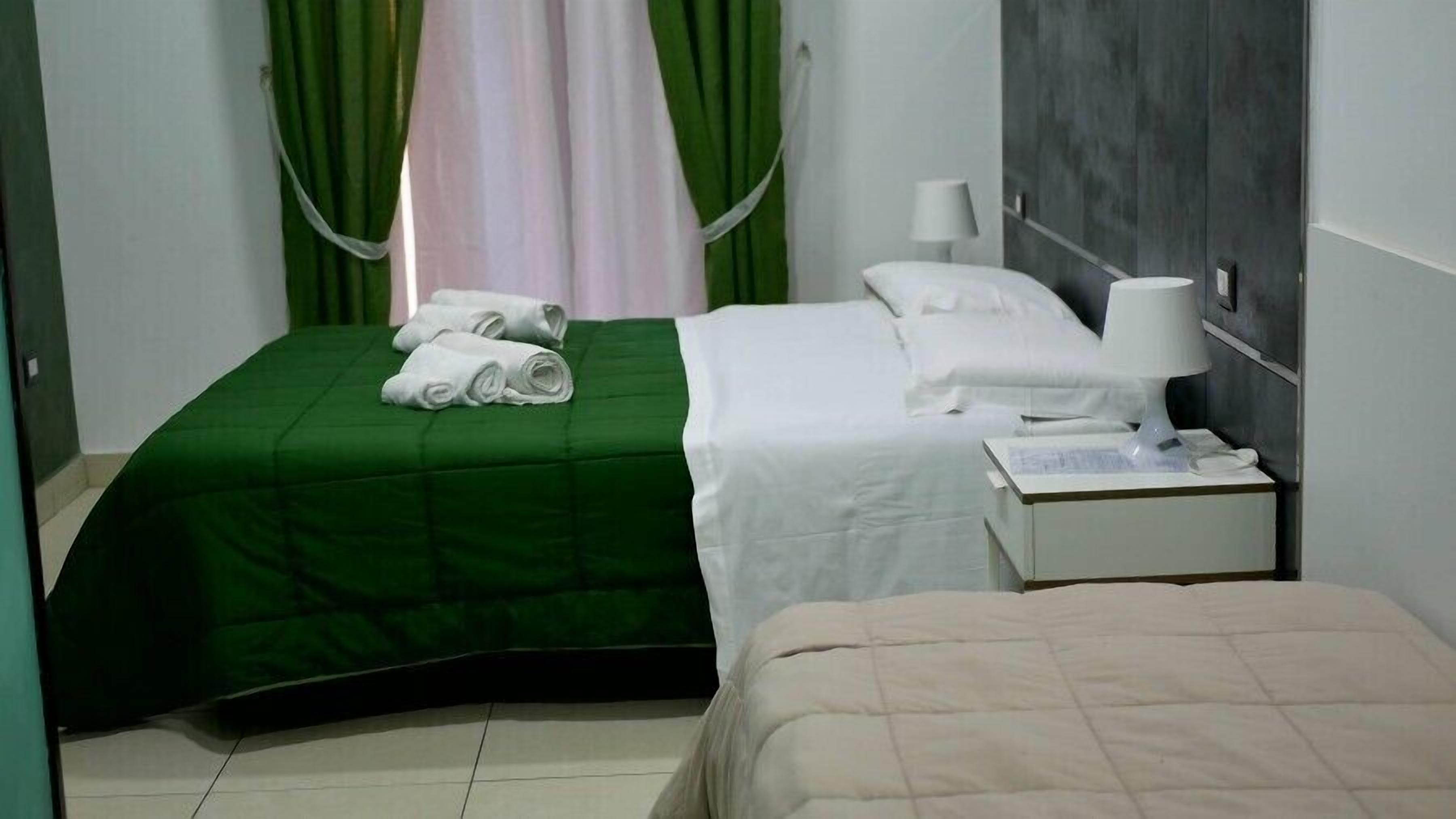 Une chambre d’hôtel avec deux lits, l’un recouvert d’une couvre-lit verte et l’autre d’une couvre-lit beige. Sur le lit recouvert de la couvre-lit verte, on trouve des serviettes enroulées. Il y a une table de chevet avec une lampe, et une petite table de nuit avec un téléphone et une boîte.