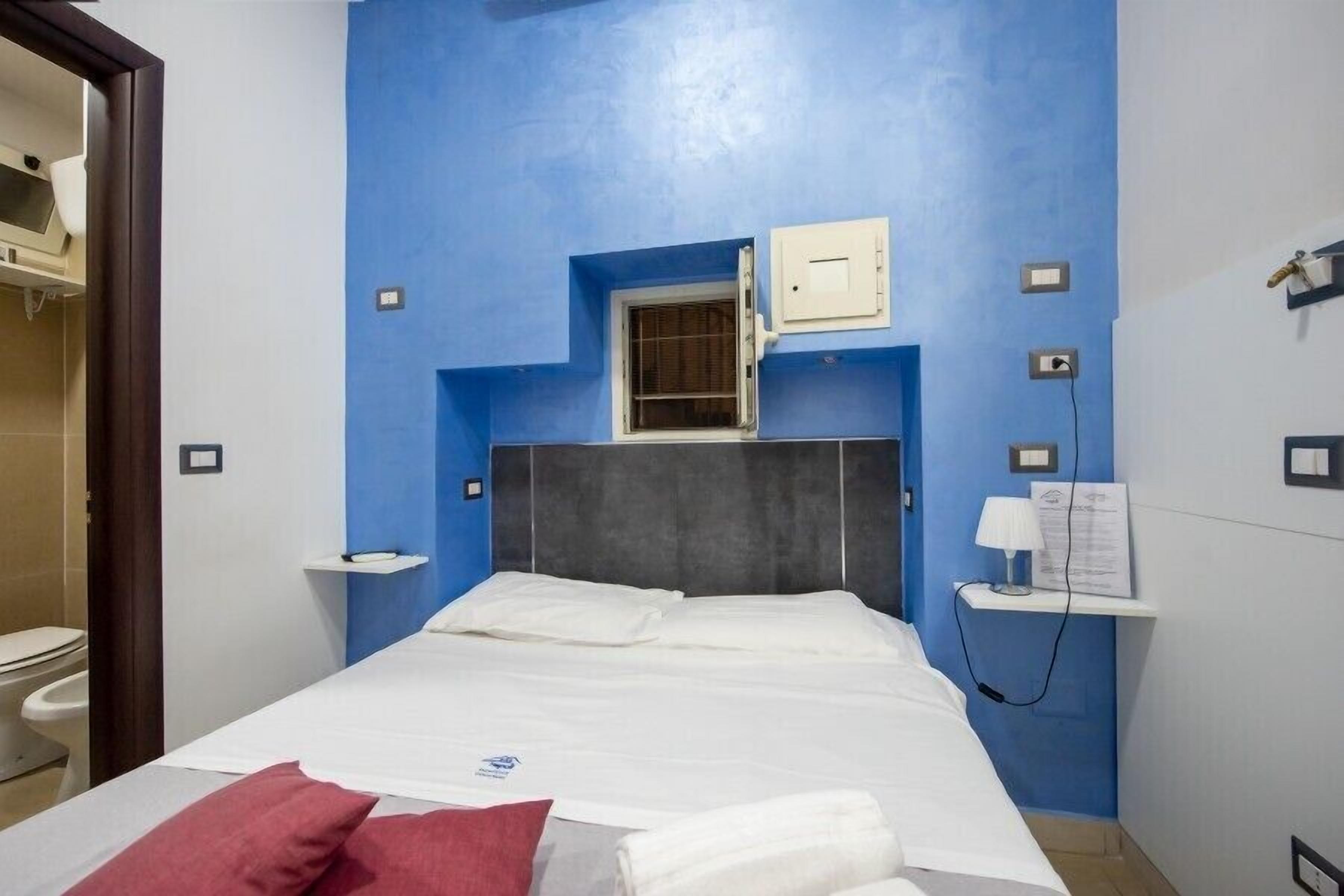 A hotel room with a bed, a window, a wall-mounted shelf with a lamp, and a small cabinet.