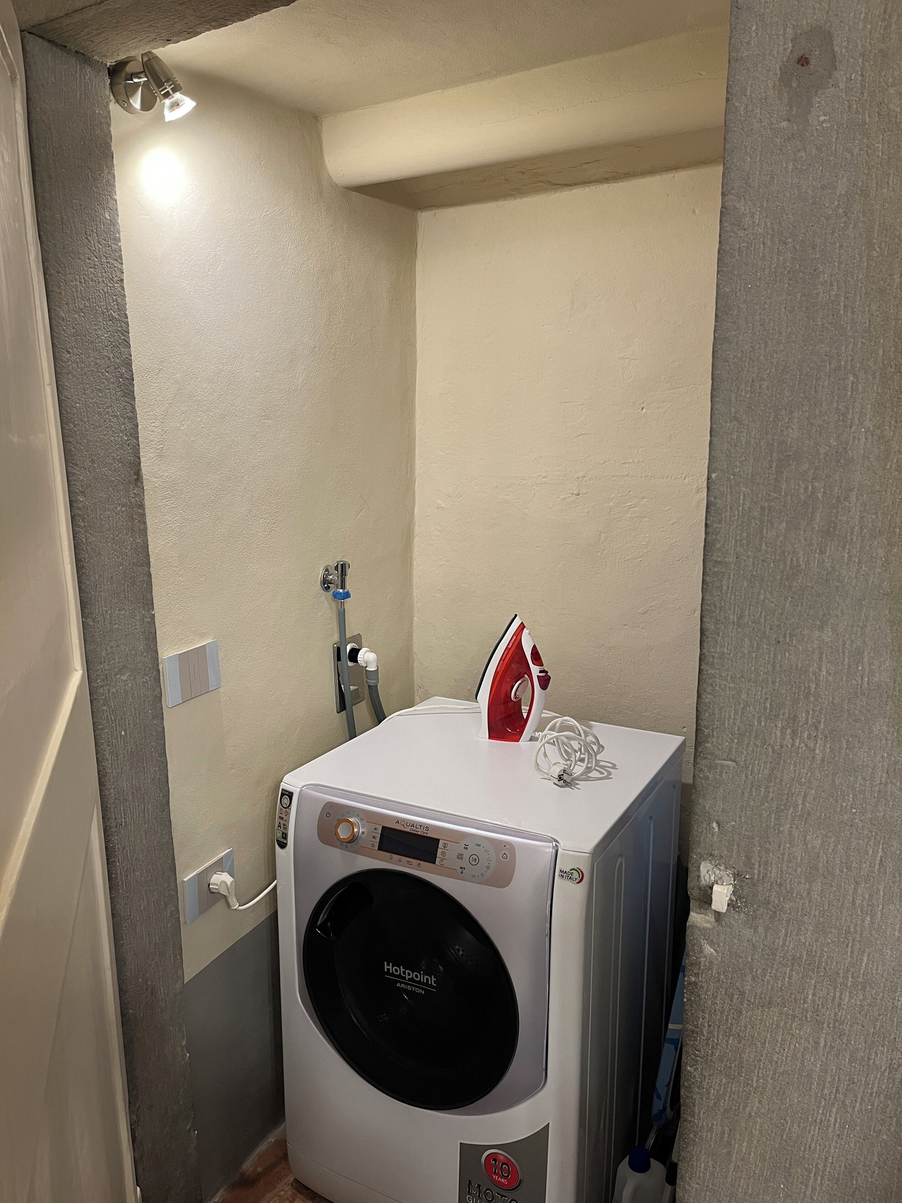 Un lave-linge compact et blanc doté d’un écran numérique, un fer à repasser rouge et blanc, et un radiateur fixé au mur dans une petite buanderie fermée.