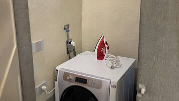 Un lave-linge compact et blanc doté d’un écran numérique, un fer à repasser rouge et blanc, et un radiateur fixé au mur dans une petite buanderie fermée.