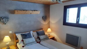 A double bed with white bedding and multiple pillows, two bedside lamps, a wooden headboard, a wooden shelf above the bed, and a window with blinds.