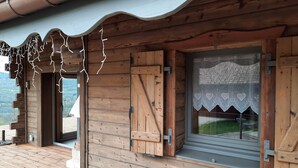 A wooden cabin with a wooden deck, a window with lace curtains, and a view of the landscape.