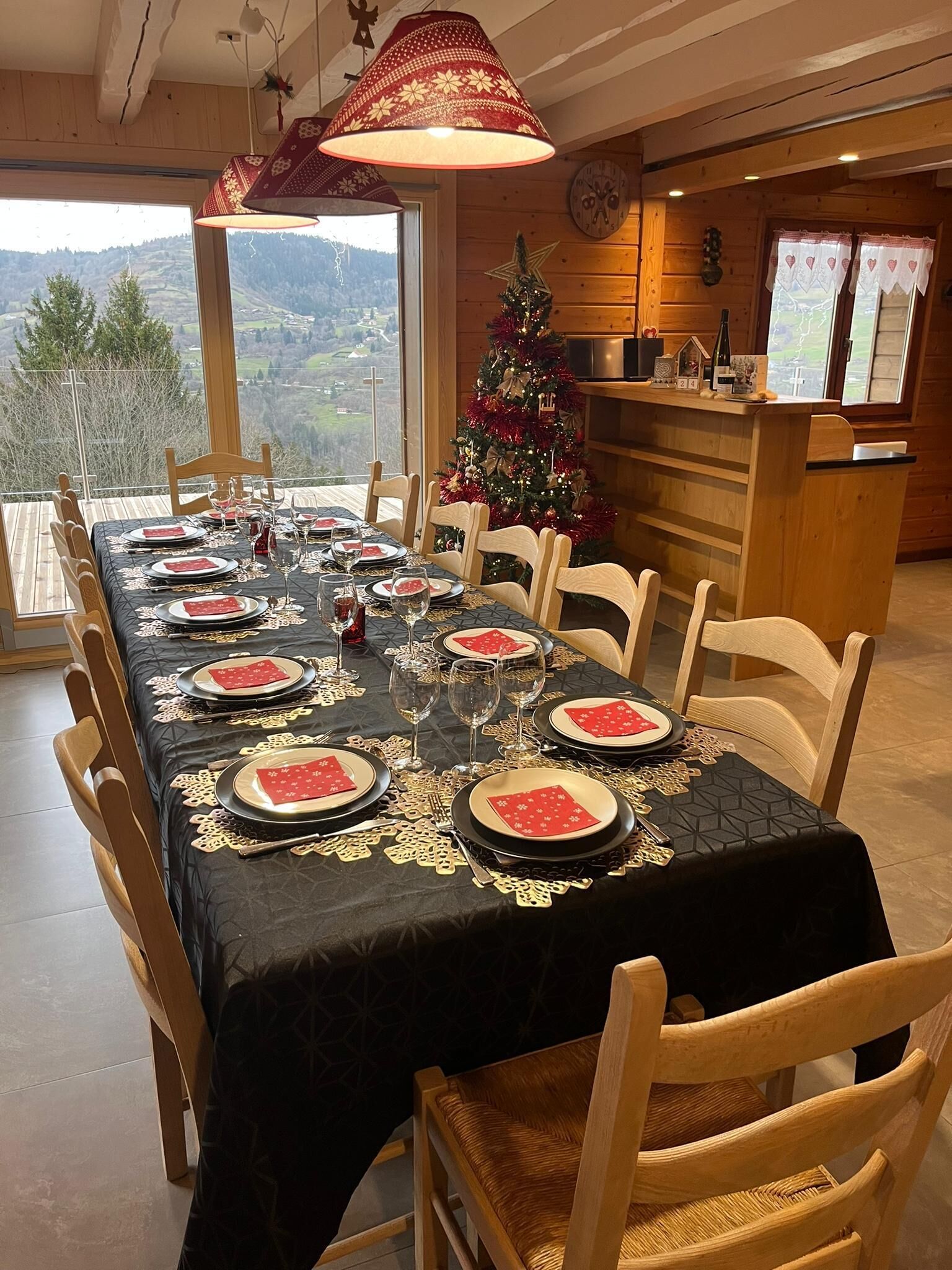 A dining table set for six with a black tablecloth and red napkins, a Christmas tree, and a view of the mountains.
