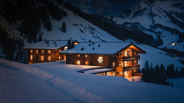 Un grand chalet en bois à plusieurs étages, dont le toit est recouvert de neige, entouré de montagnes et d’arbres.