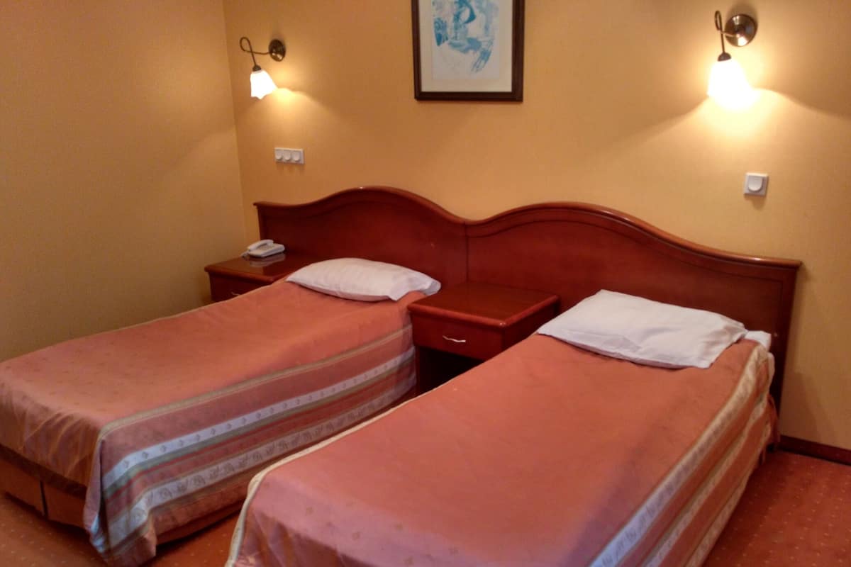 A hotel room with two single beds, wooden headboards, bedside tables with lamps, and a framed artwork on the wall.