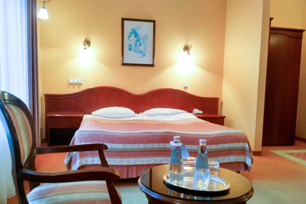 A hotel room with a bed, a chair, a bedside table with a tray of water bottles, and a framed picture on the wall.