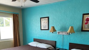 Hotellihuone, jossa on kaksi sänkyä, kattotuuletin, ikkuna verhoilla ja seinään kiinnitetyt lamput.