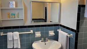Ein Badezimmer mit einem weißen Waschbecken, einer Toilette und einem Spiegel über dem Waschbecken. Es gibt Regale mit Toilettenartikeln und Handtüchern, die an der Wand hängen.
