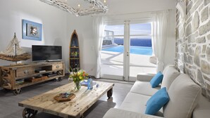 A living room with a television, wooden coffee table, and a sofa with blue cushions. There is a stone wall and a view of the ocean through a glass door. - MYKONOS CALYPSO SUNSET LUXURY VILLA POOL-HOT TUB (MYKONOS)