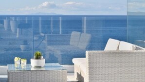 A balcony with a white wicker sofa and armchairs, a glass table with a potted plant and two glasses of water, and a view of the ocean. - MYKONOS CALYPSO SUNSET LUXURY VILLA POOL-HOT TUB (MYKONOS)