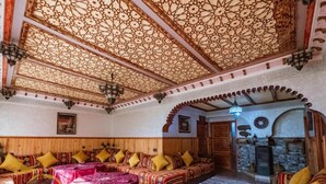 A traditional interior with a wooden ceiling, a stone fireplace, and a seating area with patterned cushions. - Mount Toubkal Lodge (Asni)