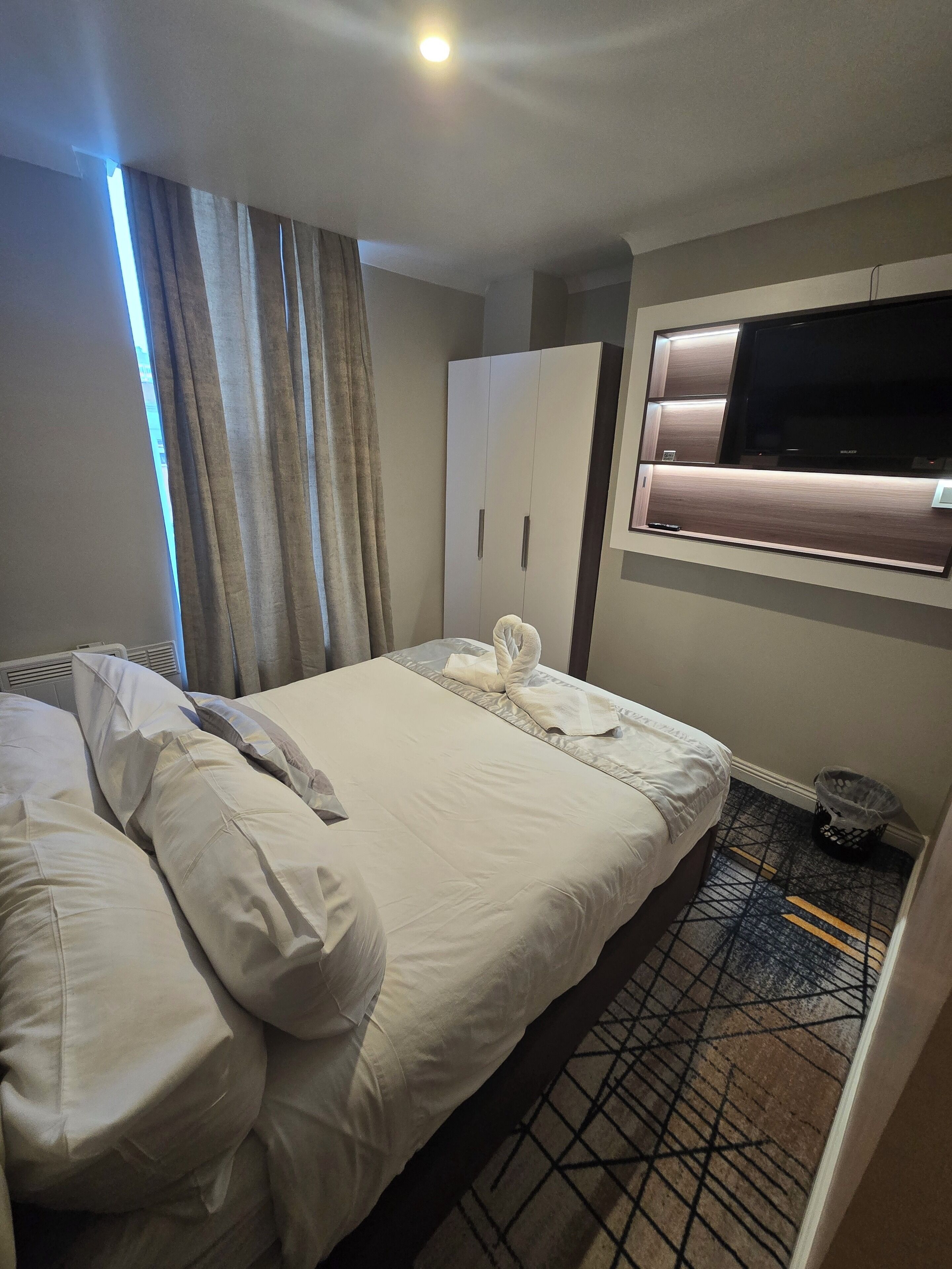 A hotel room with a bed, a wardrobe, a TV mounted on the wall, and a trash bin.