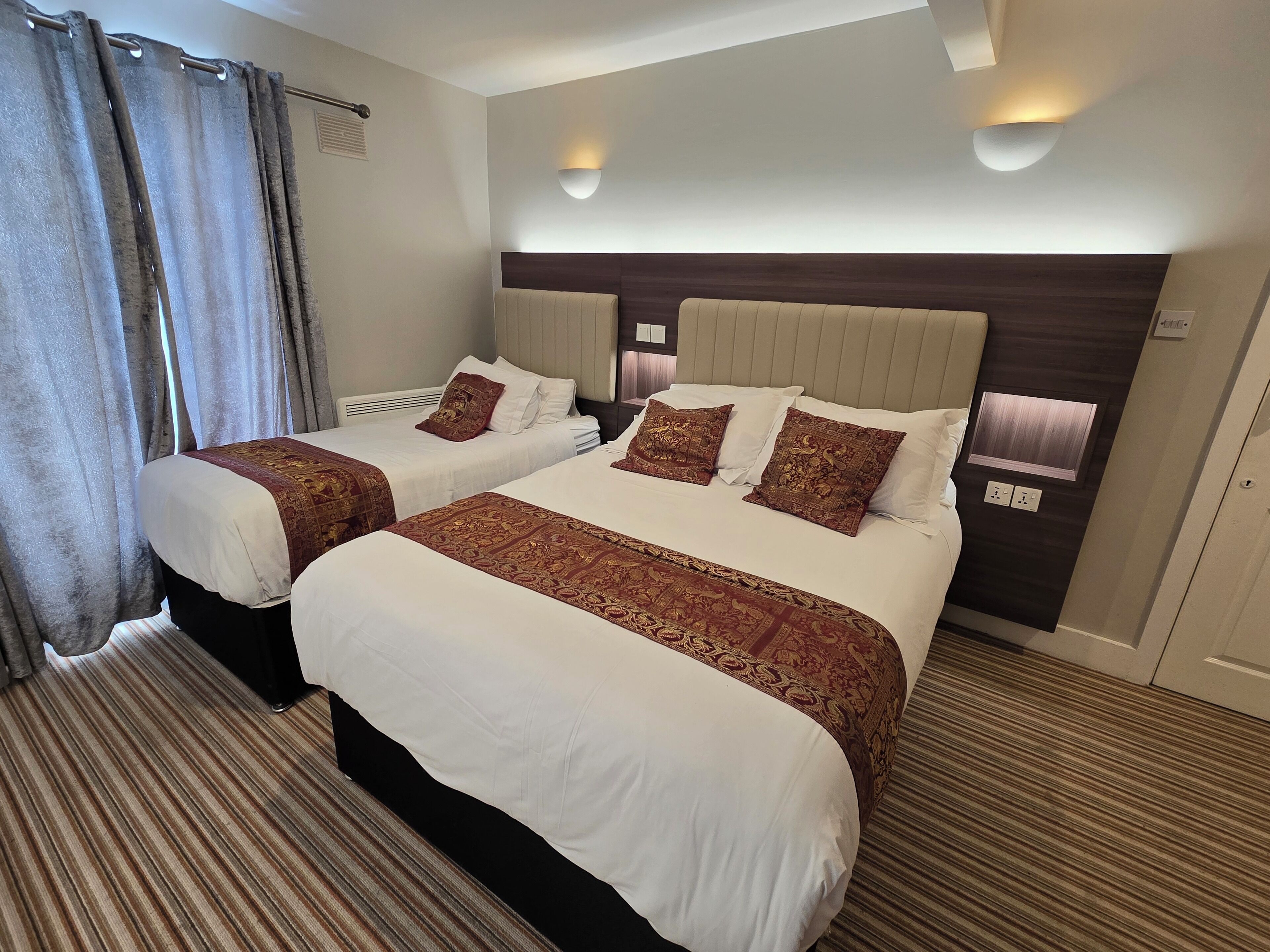 A hotel room with two beds, each with a headboard, a large window with curtains, and a wall-mounted light fixture.