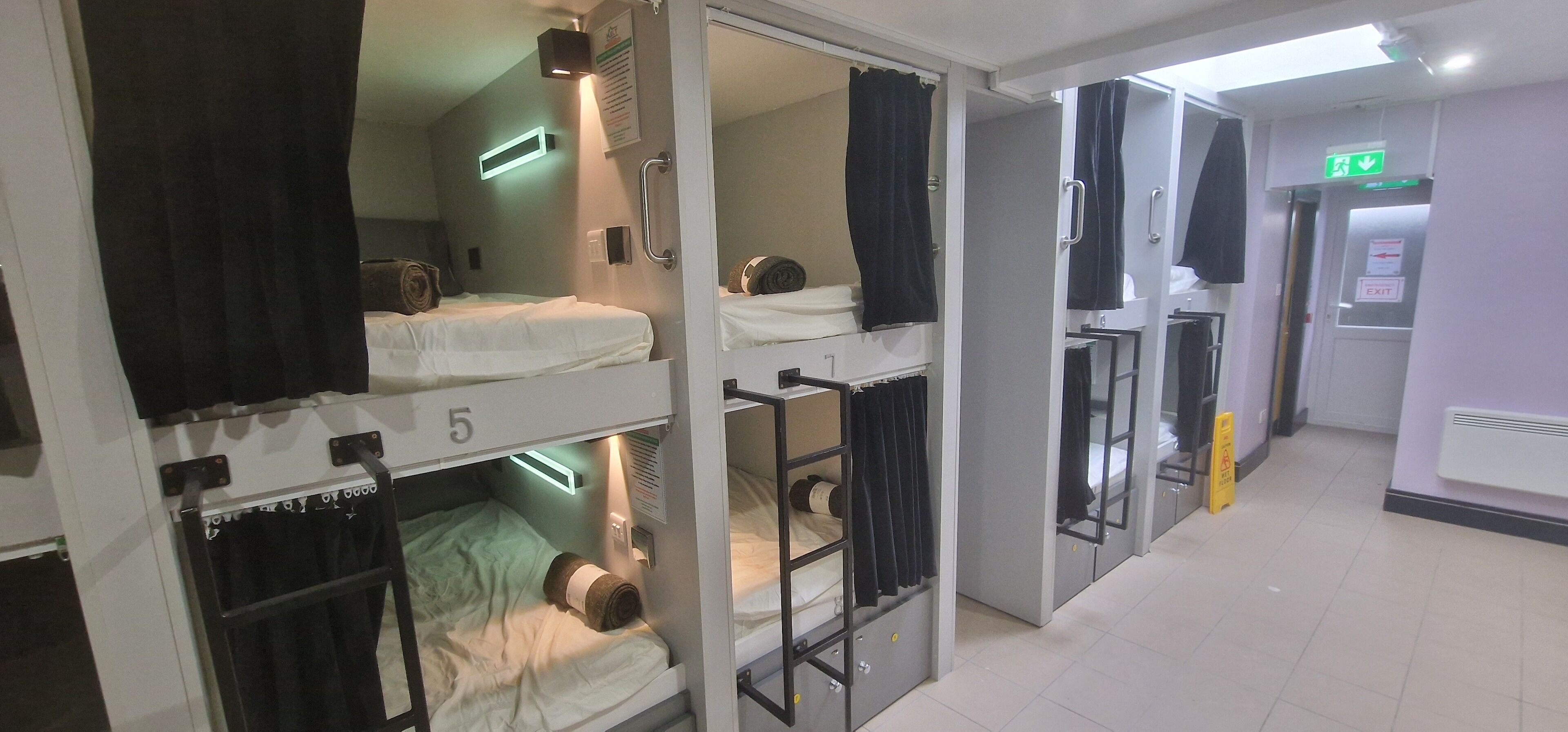 A hotel room with bunk beds, a bathroom with a shower, and a hallway with an exit sign.