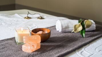 A bathroom with a white towel, a wooden bowl with pink bath salts, a lit candle, and a rolled towel on a textured bath mat.