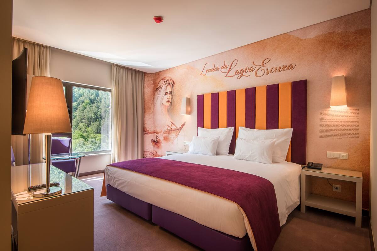 Une chambre d’hôtel avec un grand lit, un bureau avec une lampe, une fenêtre avec des rideaux et un mur orné d’une fresque et d’un texte.