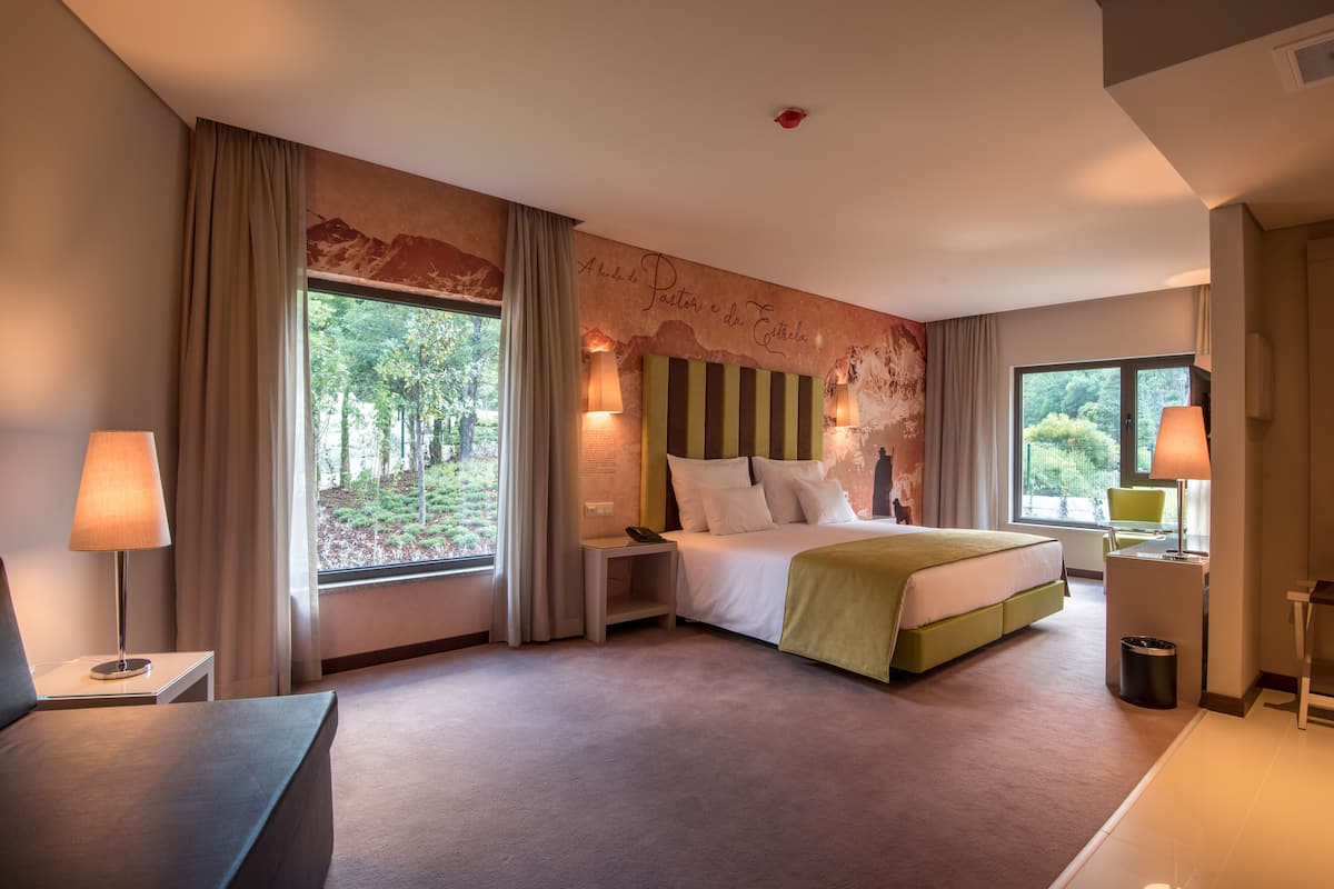 Une chambre d’hôtel avec un grand lit, deux tables de chevet, un bureau et une vue sur la verdure par les fenêtres.