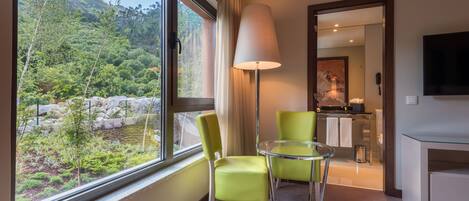 A hotel room with a large window offering a view of a mountainous landscape, a glass table with two chairs, a lamp, and a flat-screen TV mounted on the wall.