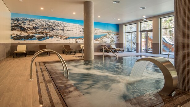 A modern indoor pool with a waterfall feature, lounge chairs, and a mural of a snowy landscape. - Vila Galé Collection Serra da Estrela Hotel (Manteigas)